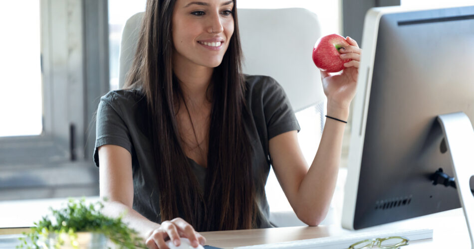 Shot of smiling young business woman eating a red apple while working with computer in the office.