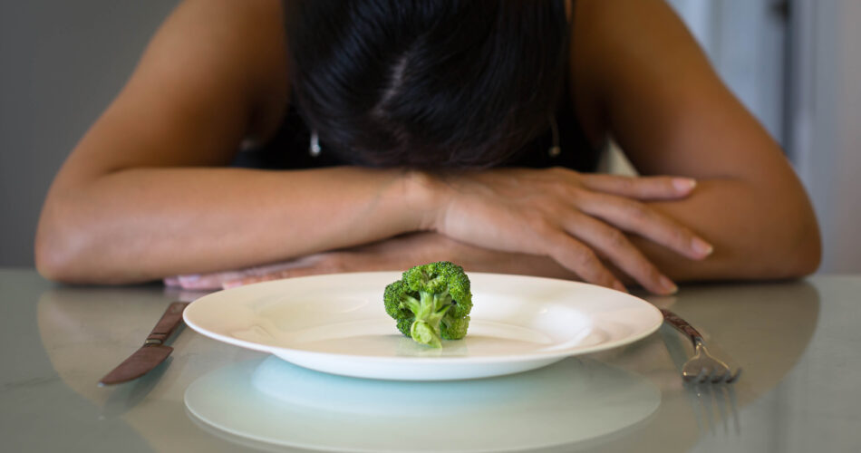 A woman sitting in front of a plate with a small portion of vegatables, looking miserable and hungry.