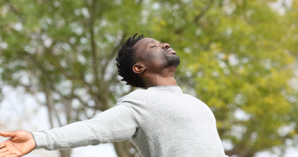 Black man breathing fresh air stretching arms in a park with a green tree in the background