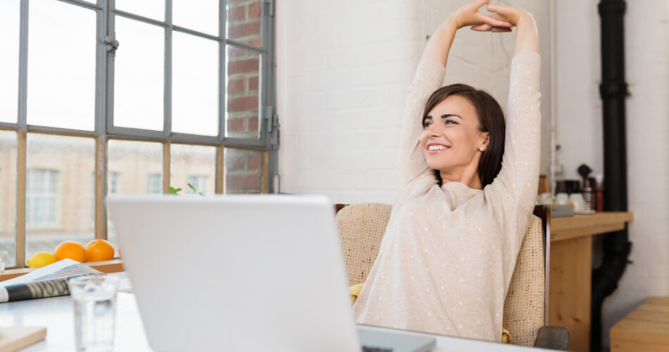 Happy relaxed young woman sitting in her kitchen with a laptop in front of her stretching her arms above her head and looking out of the window with a smile