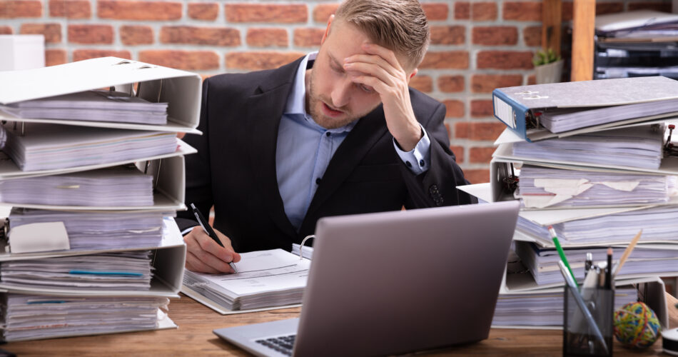 Stressful Businessman Sitting In Office With Stacked Of Files