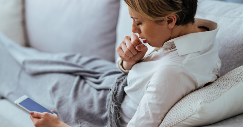 Woman having cold and flue virus and coughing while using cell phone on the sofa.