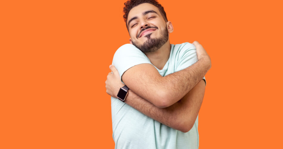 I love myself! Portrait of egoistic brunette man with beard in white t-shirt standing with closed eyes, embracing himself and smiling form pleasure and proud. indoor, isolated on orange background